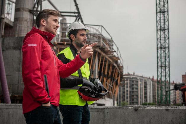 Two men, one in a Danfoss jacket and the other in a safety vest and hard hat, pointing to something on a construction site