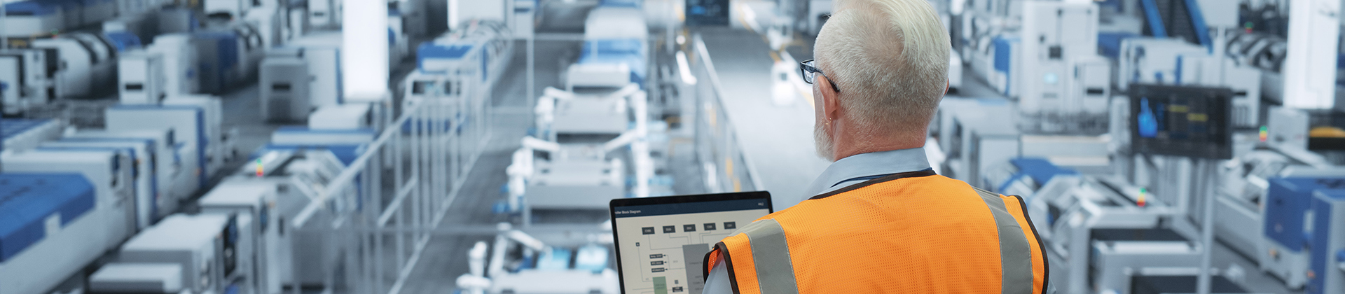 Man in a safety vest looking at his open laptop while standing above a factory floor
