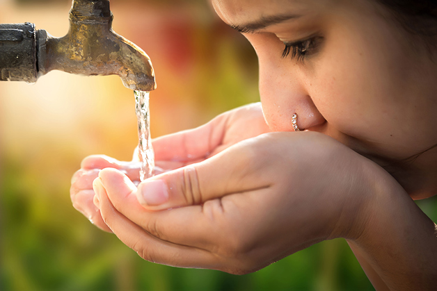 A young lady with a nose ring filling her hands with water from a weathered spigot and taking a sip.